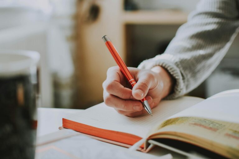 Someone writing in a small notebook on a desk with a coffee cup, in soft morning light